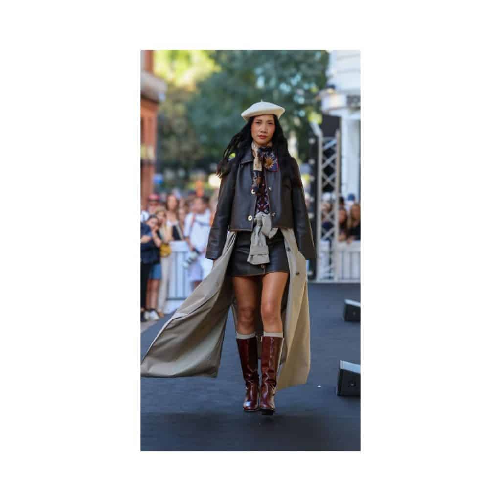 mannequin défilant sur un podium en extérieur lors d'un défilé de Galerie Lafayette, portant une tenue élégante avec manteau long, veste, bottes hautes.