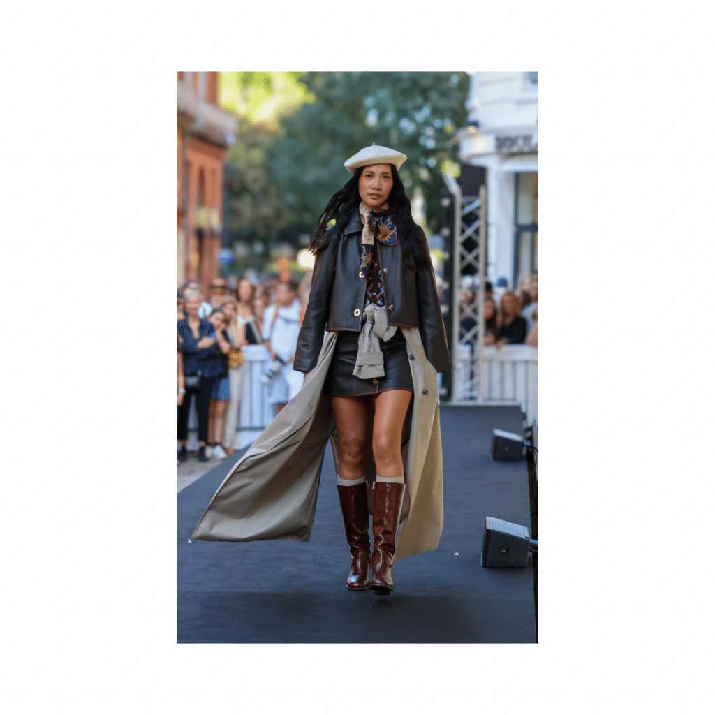 mannequin défilant sur un podium en extérieur lors d'un défilé de Galerie Lafayette, portant une tenue élégante avec manteau long, veste, bottes hautes.